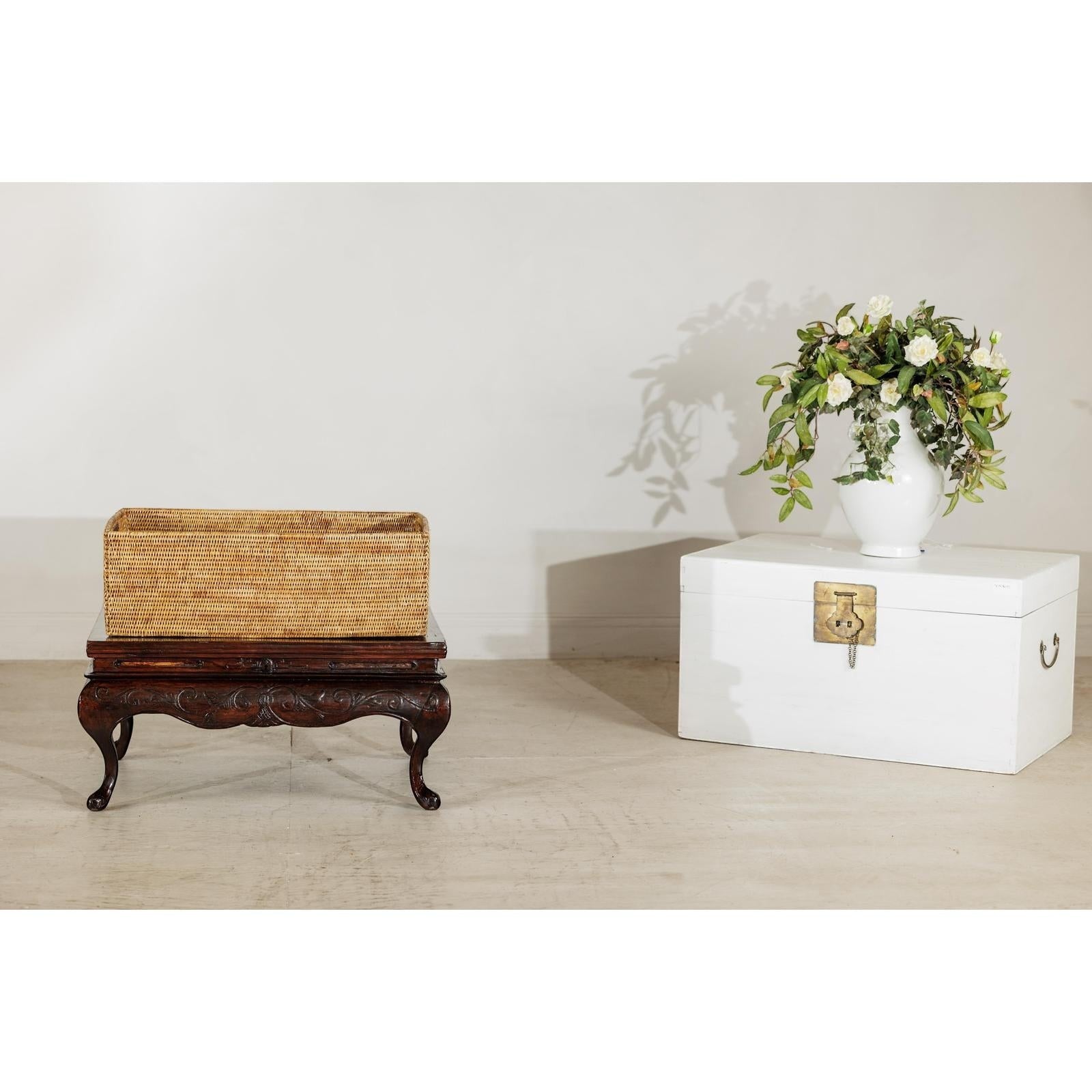 Close-up of a vintage rectangular handwoven rattan basket with side handles beside a white chest and floral arrangement.