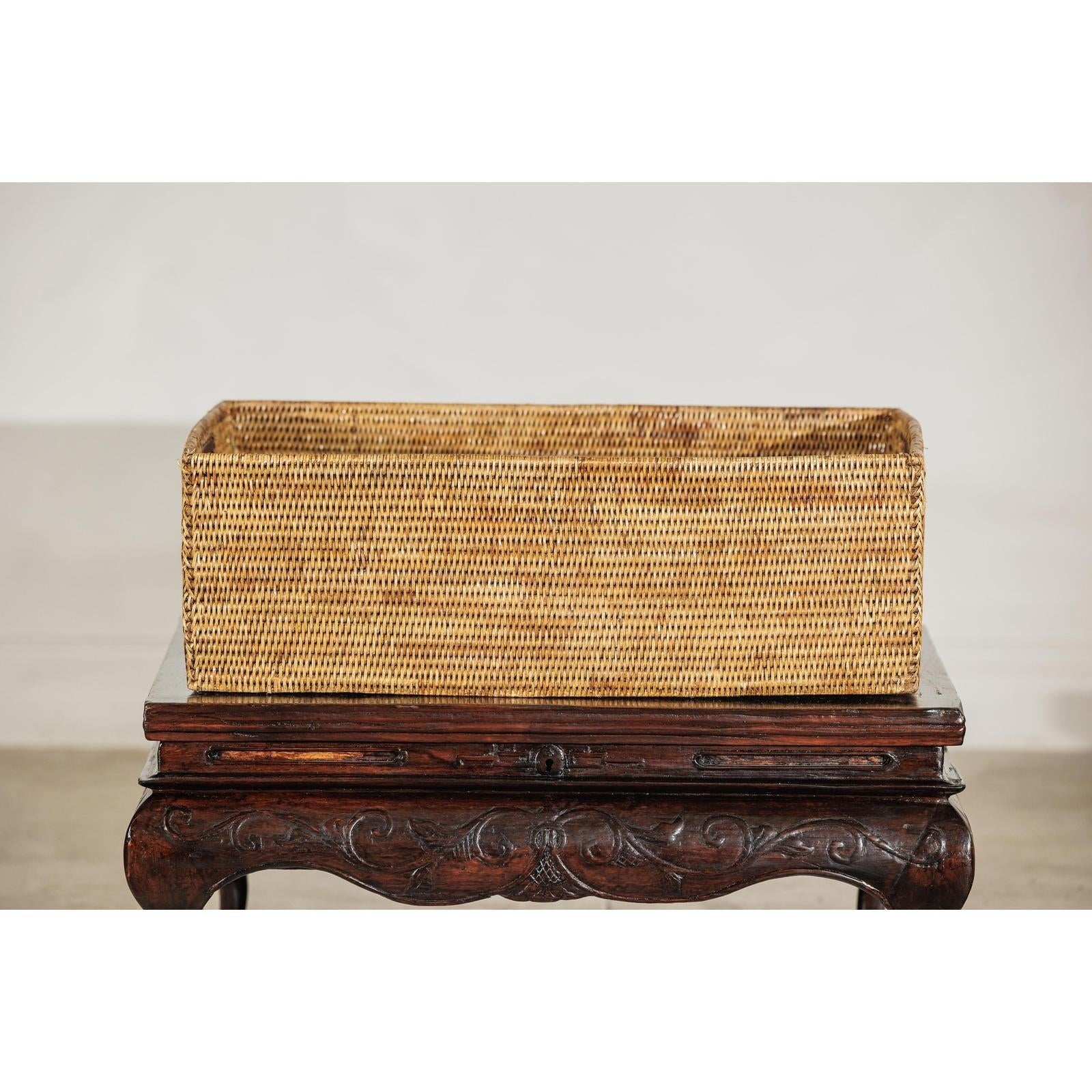 Brown handwoven rattan basket with side handles on ornate dark wooden table, plain backdrop, close-up view.