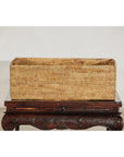 Brown handwoven rattan basket with side handles on ornate dark wooden table, plain backdrop, close-up view.