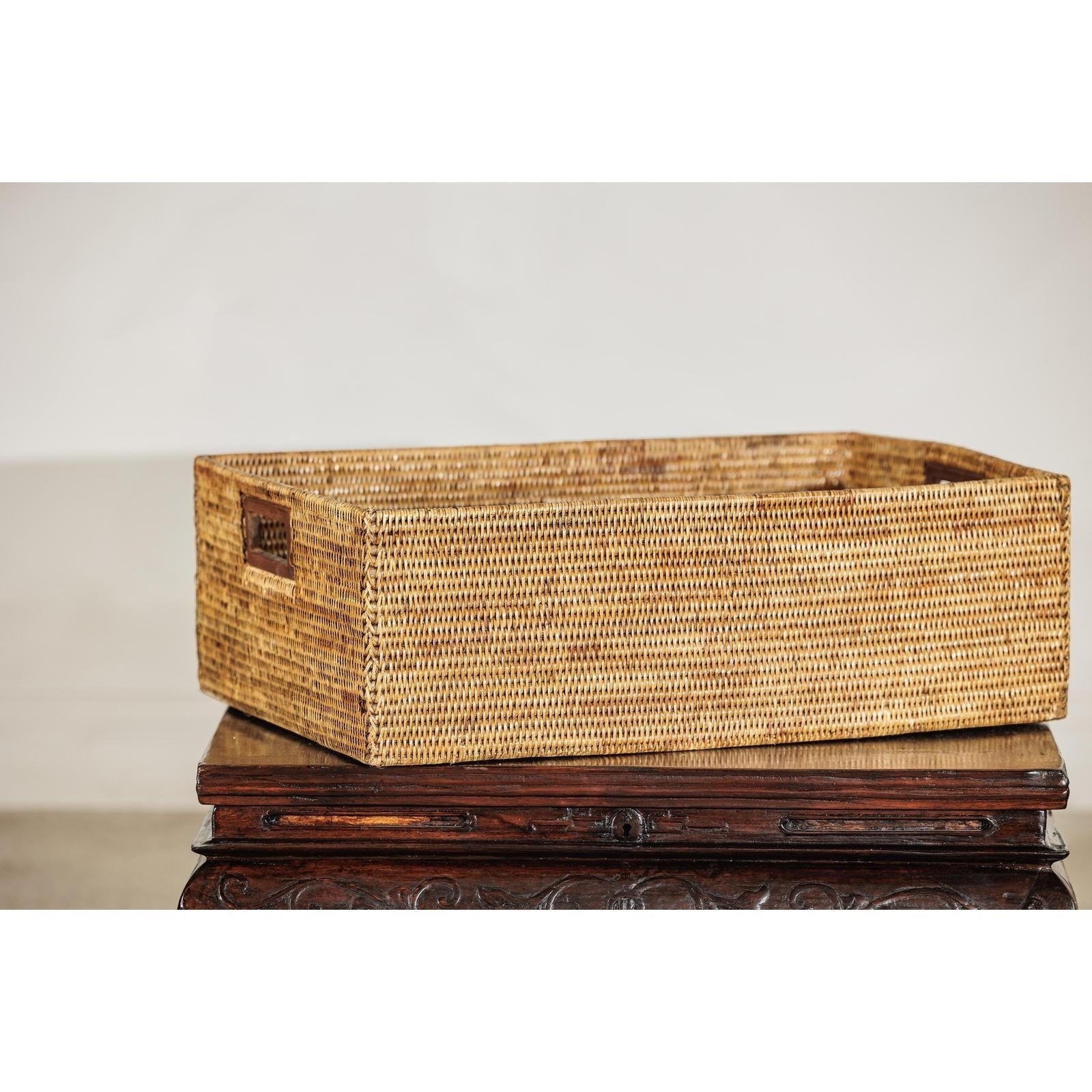 Brown handwoven rattan rectangular basket with side handles on carved wooden surface against a plain backdrop.