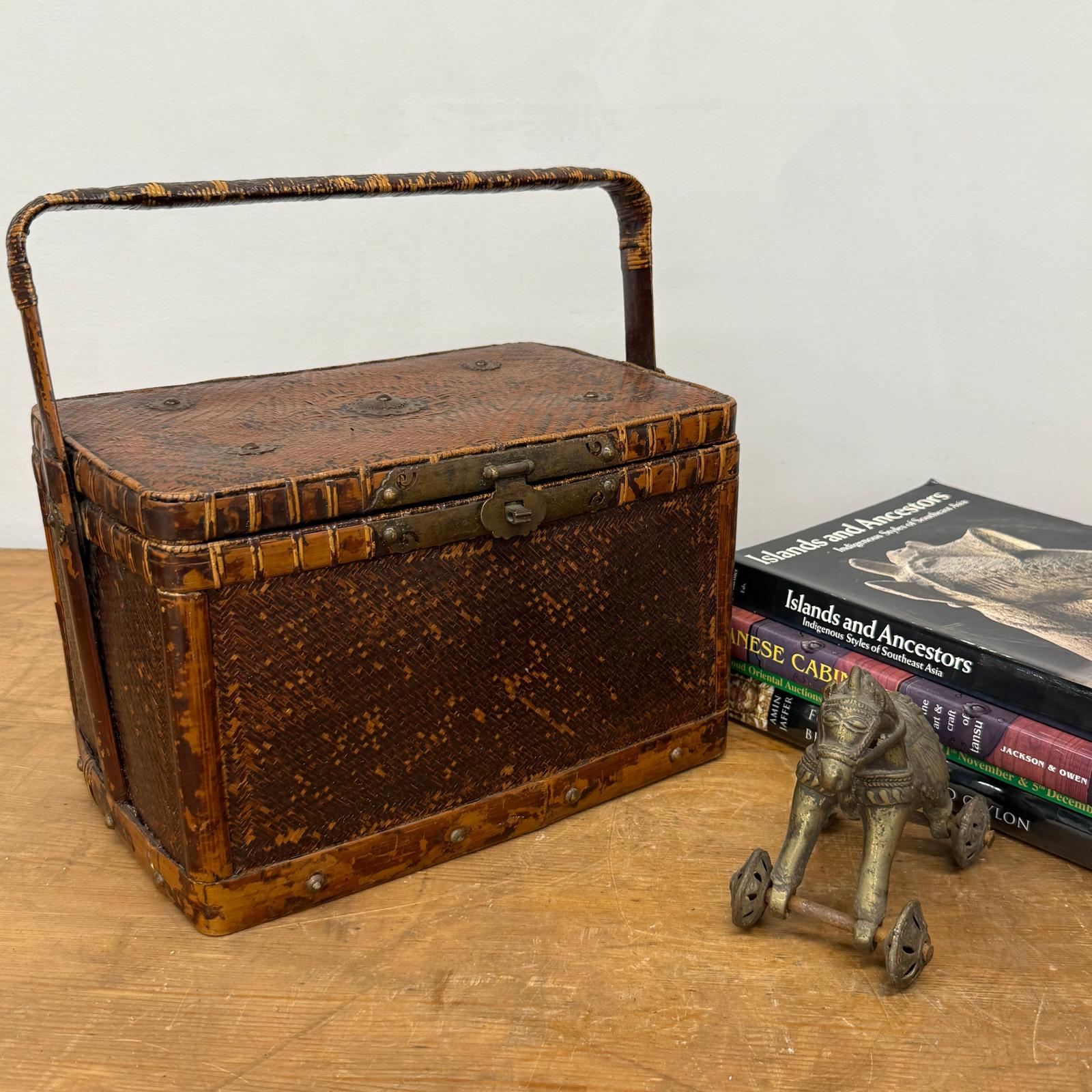The Woven Rectangular Picnic Basket with Handle &amp; Hinged Lid sits on a wooden surface beside stacked books and a small metal elephant figurine, showing off exquisite Southeast Asian craftsmanship.