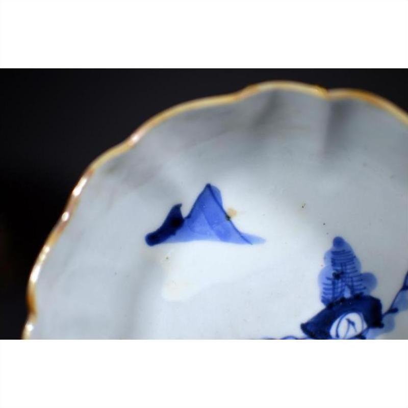 Close-up of blue and white hand painted porcelain bowl with gold rim, scalloped edge, and mountain scene.