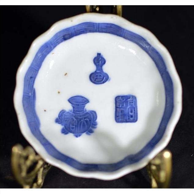 Blue and white porcelain dish, hand painted with Asian design, displayed on brass stand against dark background.