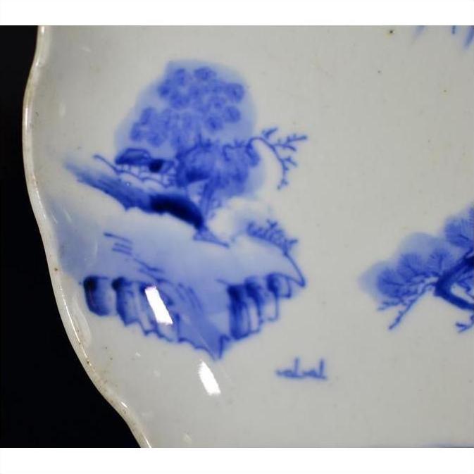 Close-up of blue and white porcelain plate with hand painted mountain landscape and willow trees, scalloped edge.