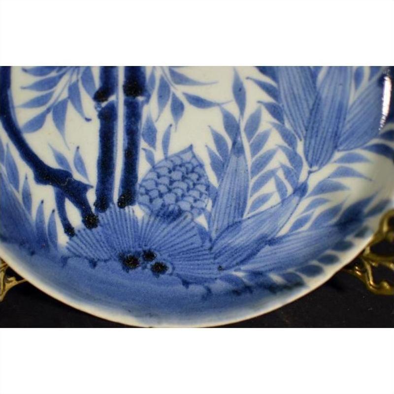 White porcelain plate with hand painted pine cone cluster and dense foliage, displayed on brass stand.