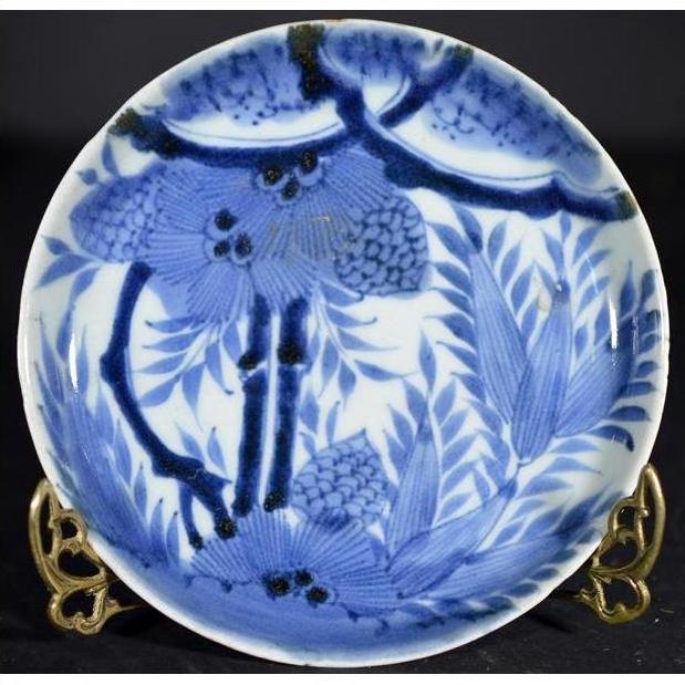 White porcelain plate with Asian design featuring pine cone cluster and dense foliage, on a decorative brass stand.