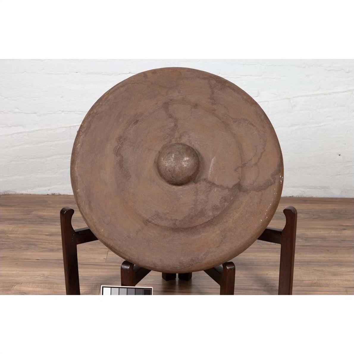Bronze Burmese temple gong with raised center and red mallet on wooden stand, against a white wall.