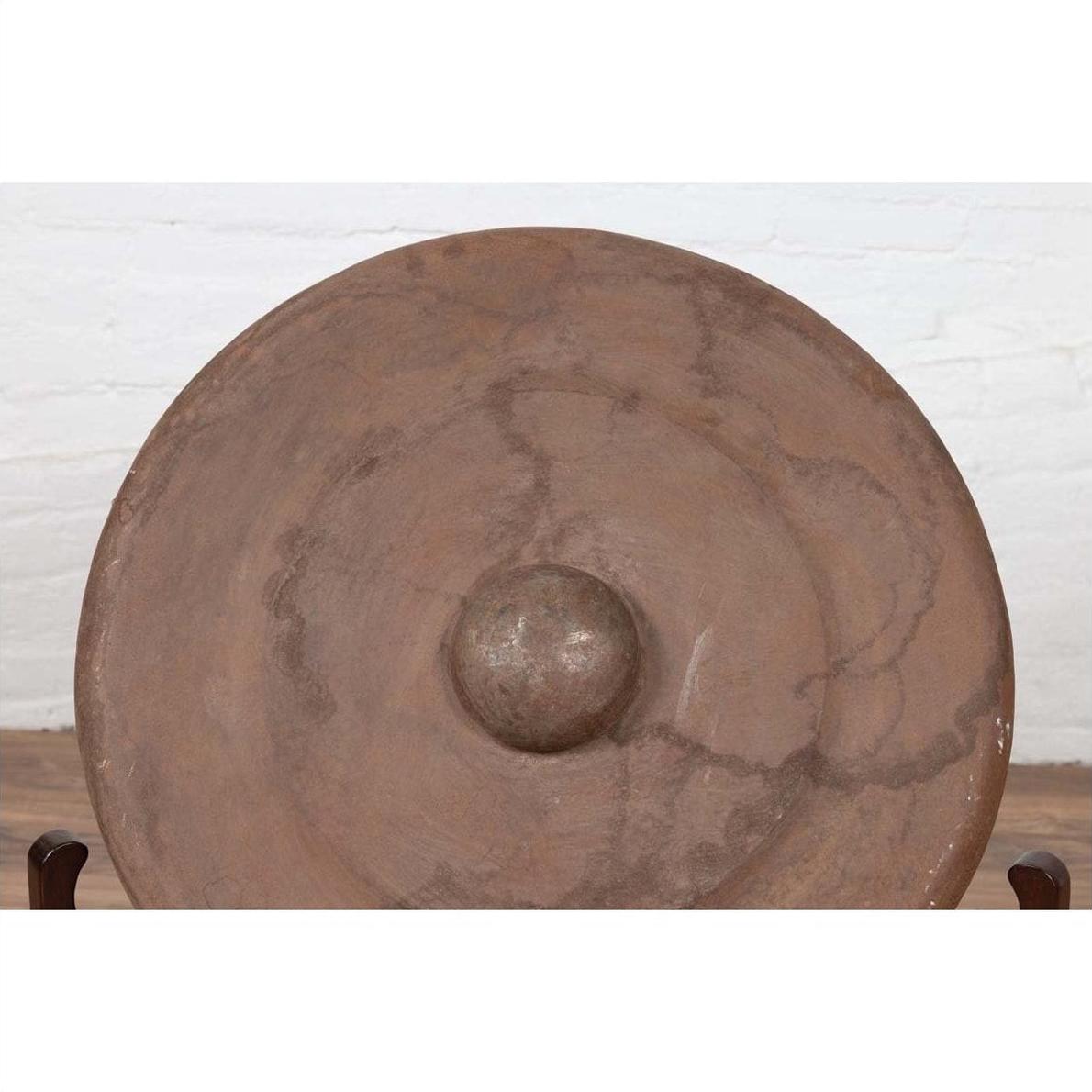 Bronze Burmese temple gong with raised center and red mallet, set against a white brick wall.