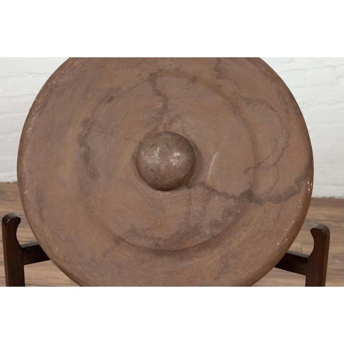 Bronze Burmese temple gong with raised center and red mallet on stand against white brick wall.