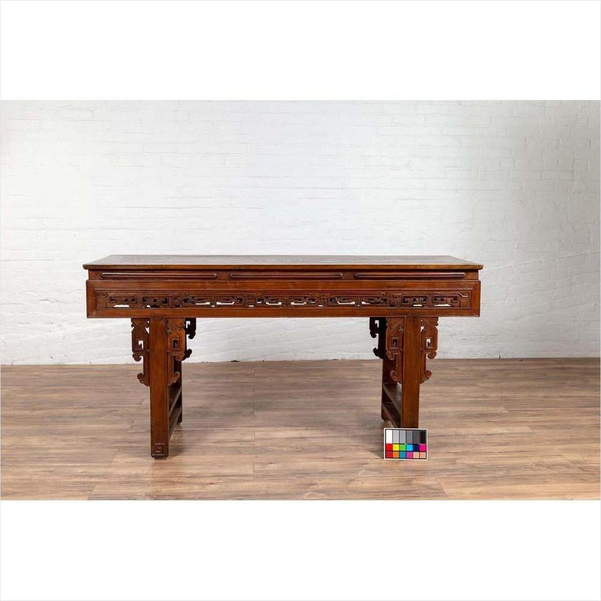 Dark wood Chinese altar console table with carved scrollwork, overhang top, on wood floor against white brick wall.