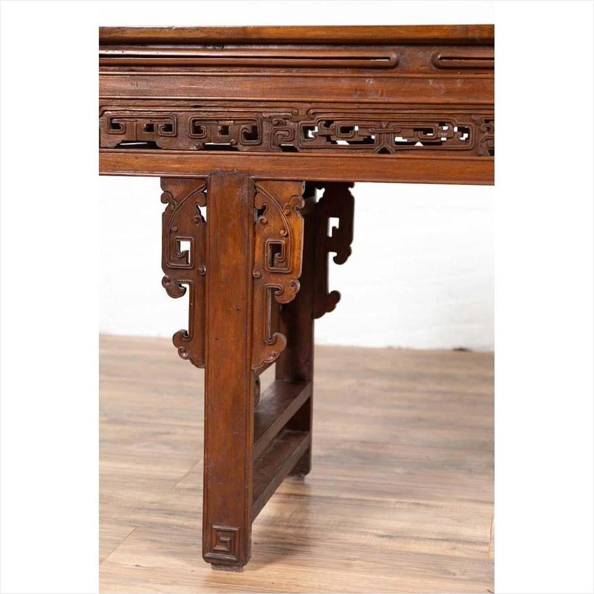 Brown wood Chinese altar console table with overhang top and open fretwork scrolls, on wooden floor by white wall.