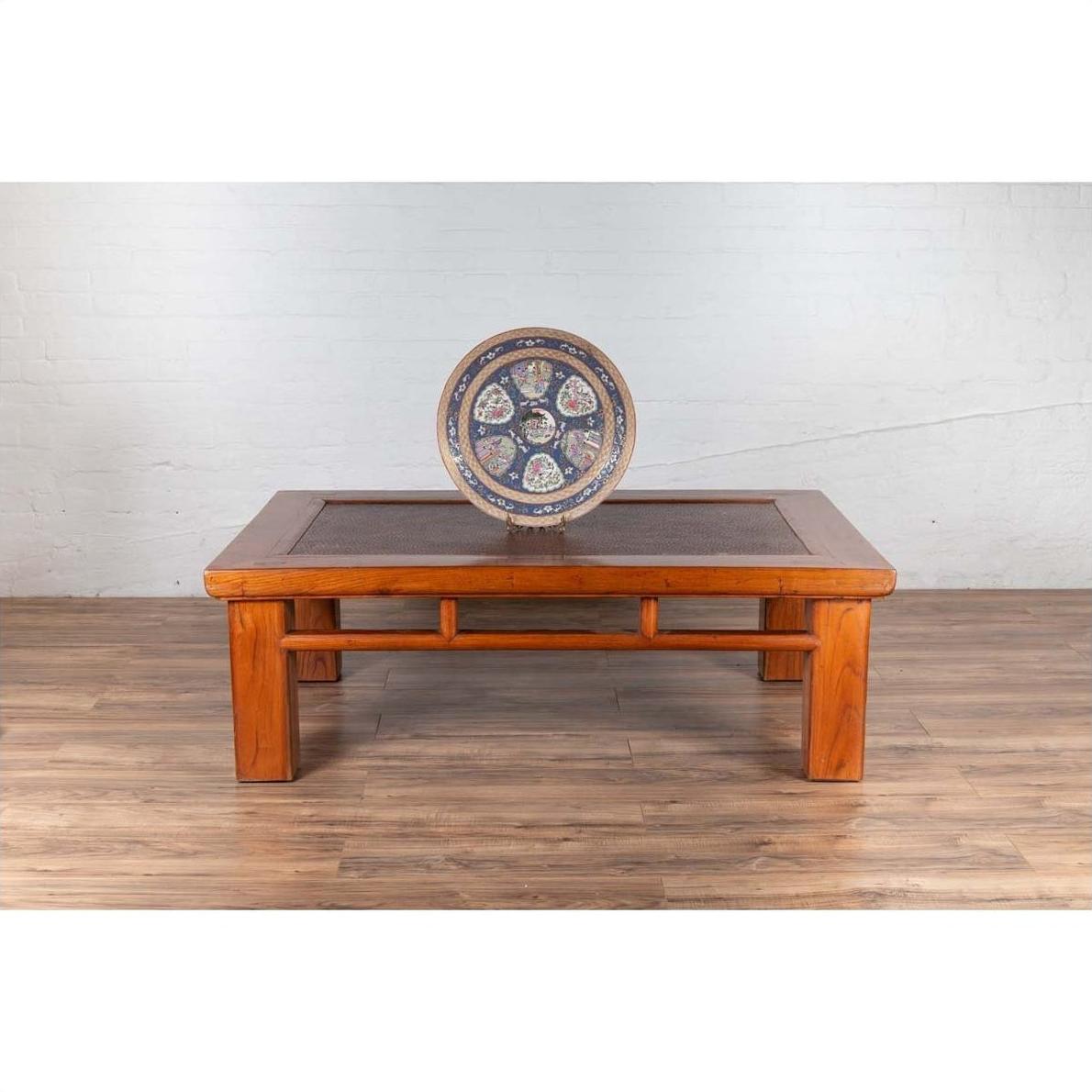 Brown Chinese elm coffee table with square legs and rattan inset top, set against a white brick wall.
