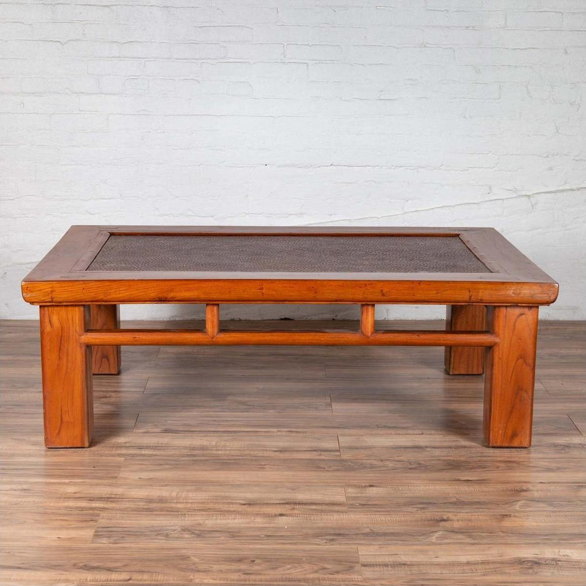 Brown Chinese elm coffee table with square legs and rattan inset top, shown on wood floor against white brick wall.