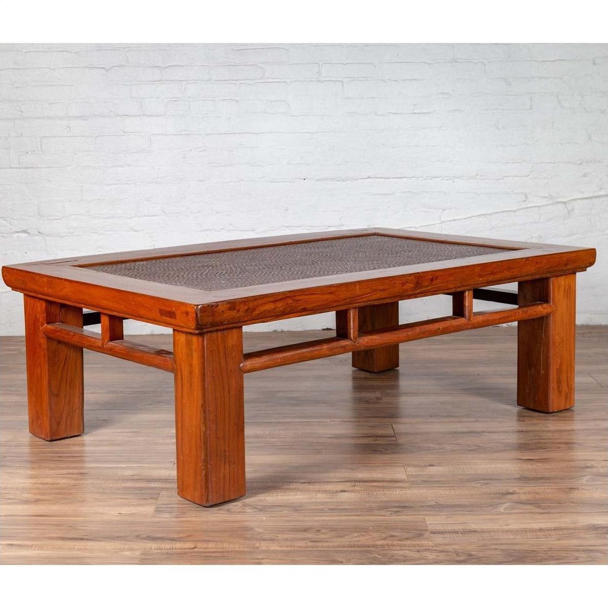 Brown Chinese elm coffee table with rattan inset top and square legs on wooden floor, white brick wall background.