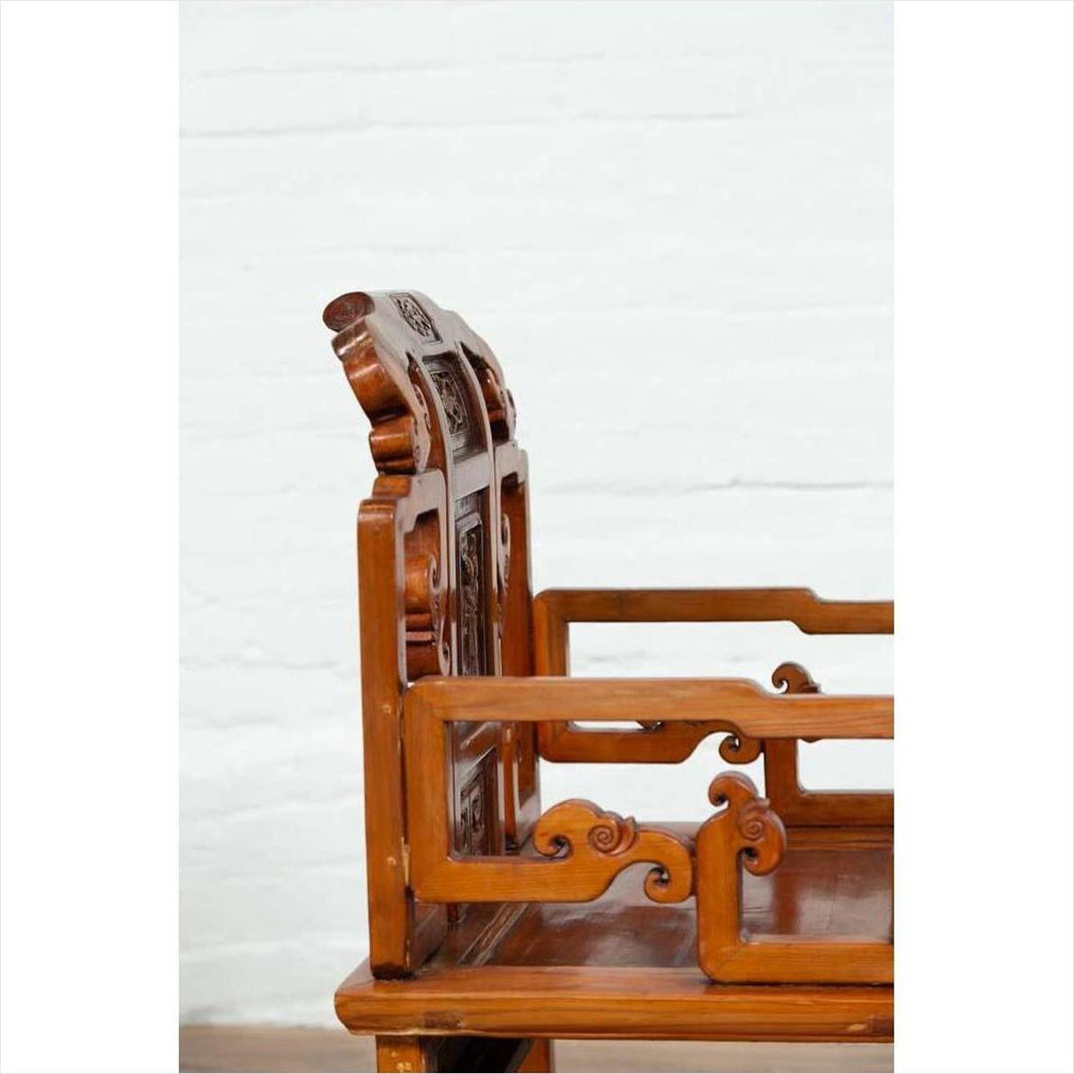 Brown wood chair with carved details on backrest and armrests, placed indoors against a white wall.