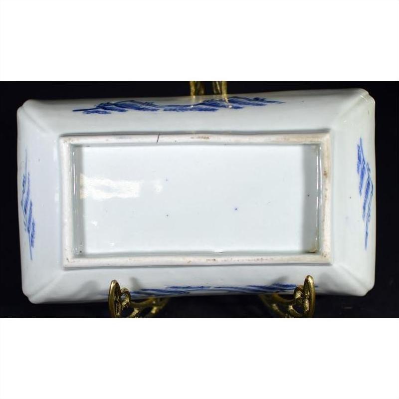 White and blue porcelain tray with landscape, pavilion, willow trees on brass stand, black background, close-up.