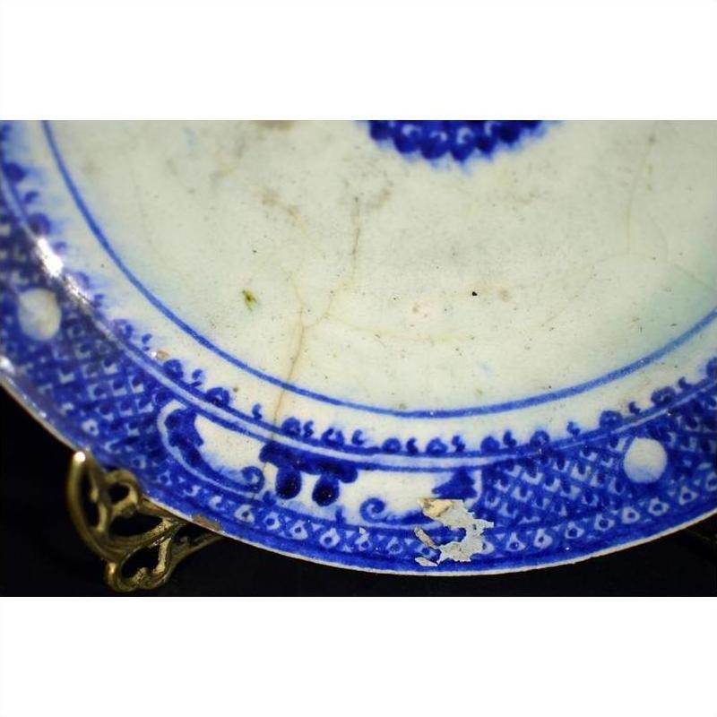 Close-up of antique ceramic plate with radial medallion, inner sunburst, loop border, visible chips and cracks.
