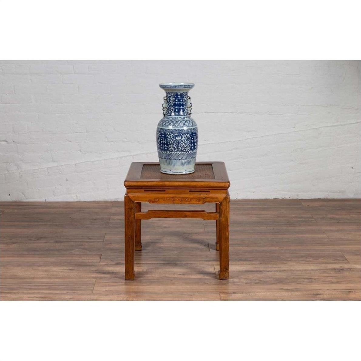 Brown elmwood Ming style side table with horsehoof legs, blue and white ceramic vase on top, against white brick wall.