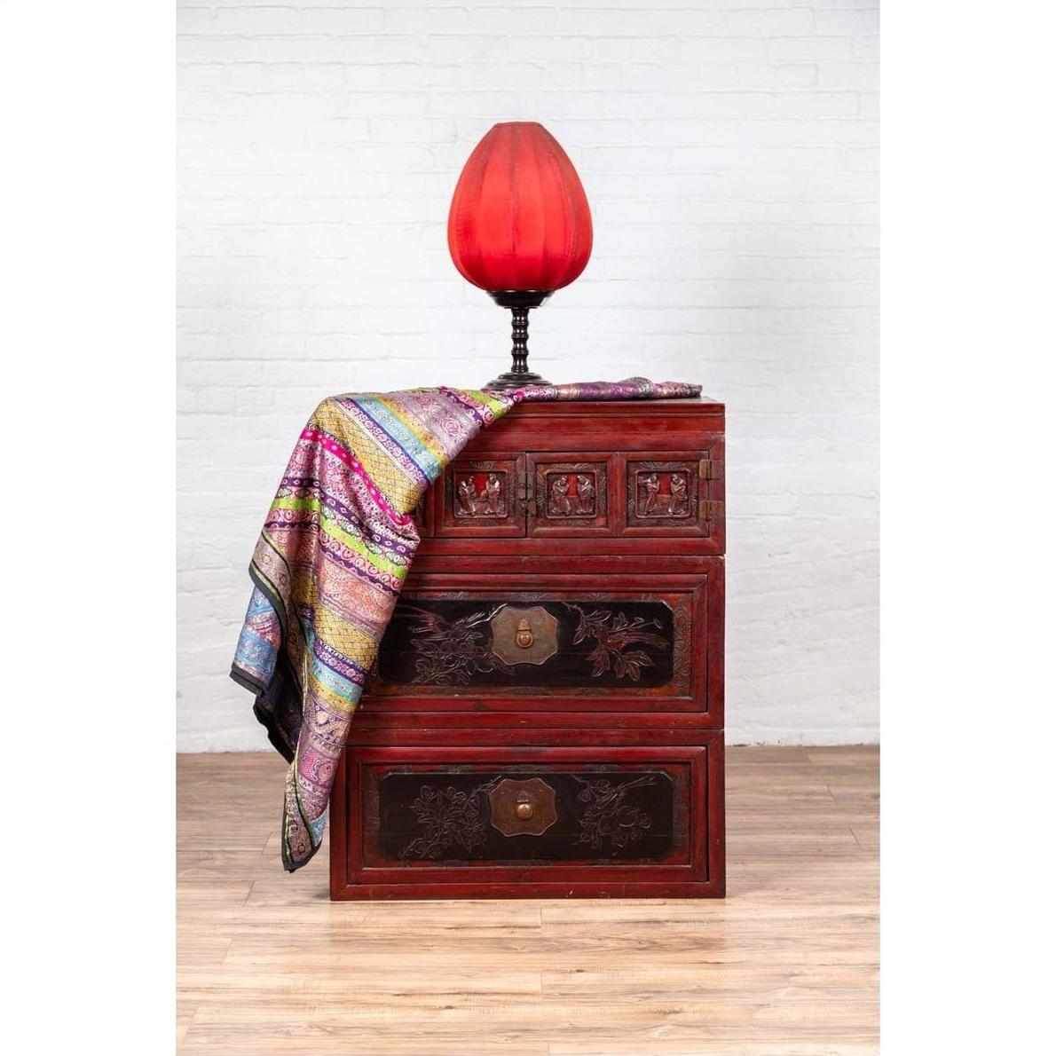 Red and black lacquered antique Chinese chest with carved figures, topped with red lamp and colorful fabric, close-up view.
