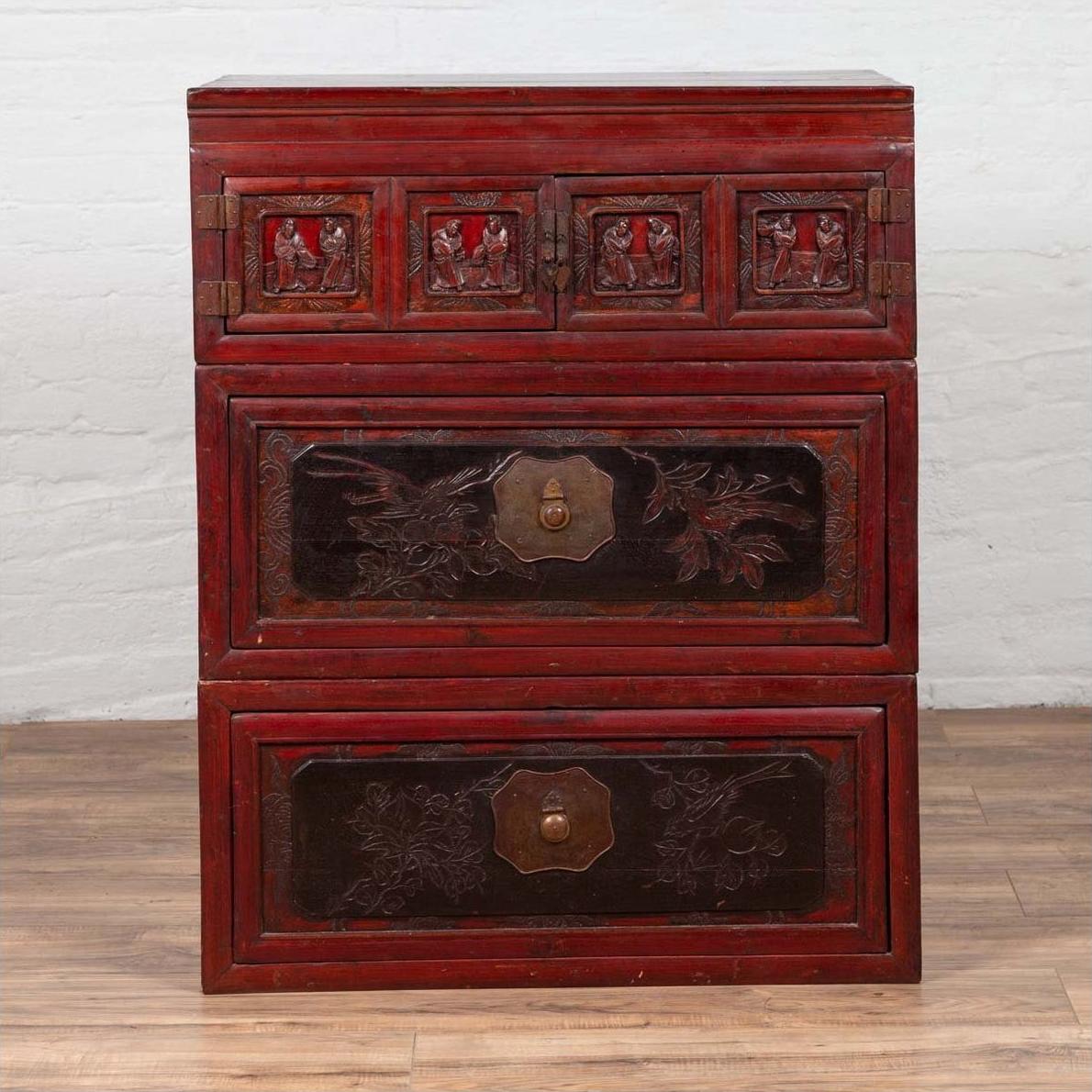 Red and black lacquered antique Chinese three-section chest with carved figures against a white wall.