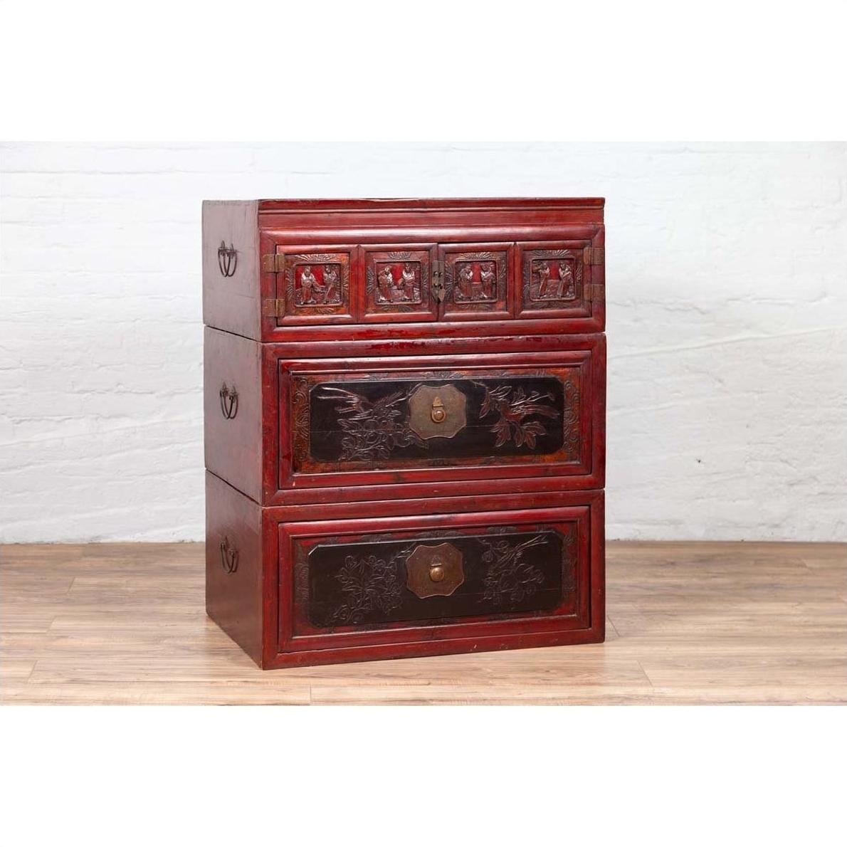 Red and black lacquered antique Chinese three-section chest with carved figures, set against a white wall.