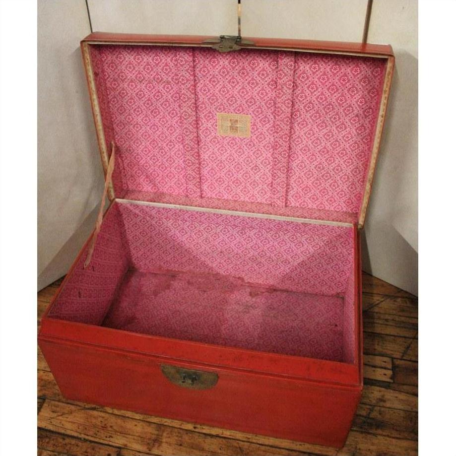 Red lacquer antique Chinese blanket chest with pink interior, open on wooden floor, light background.