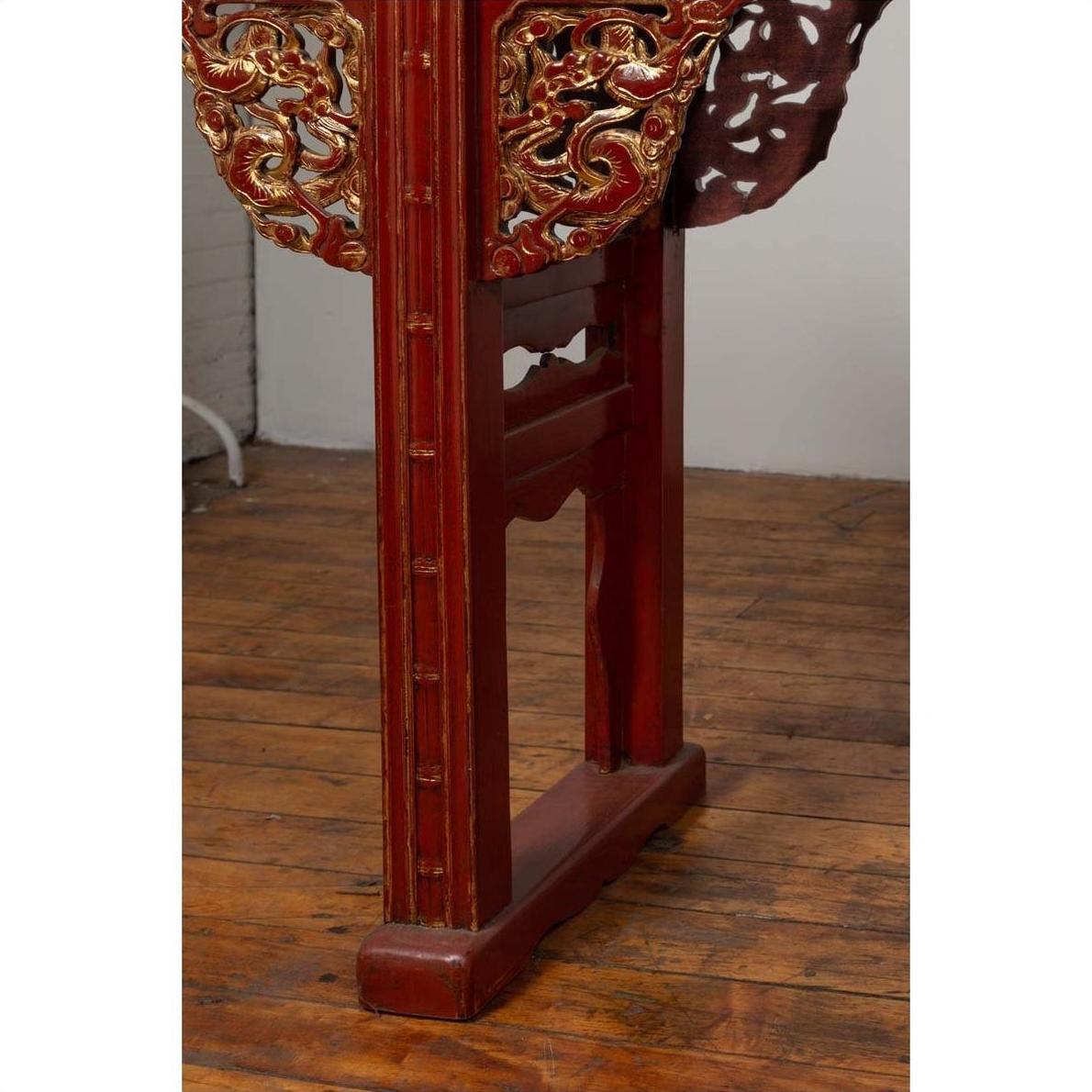 Close-up of red lacquered carved wood table leg with gold accents on a wood floor against a white wall indoors.