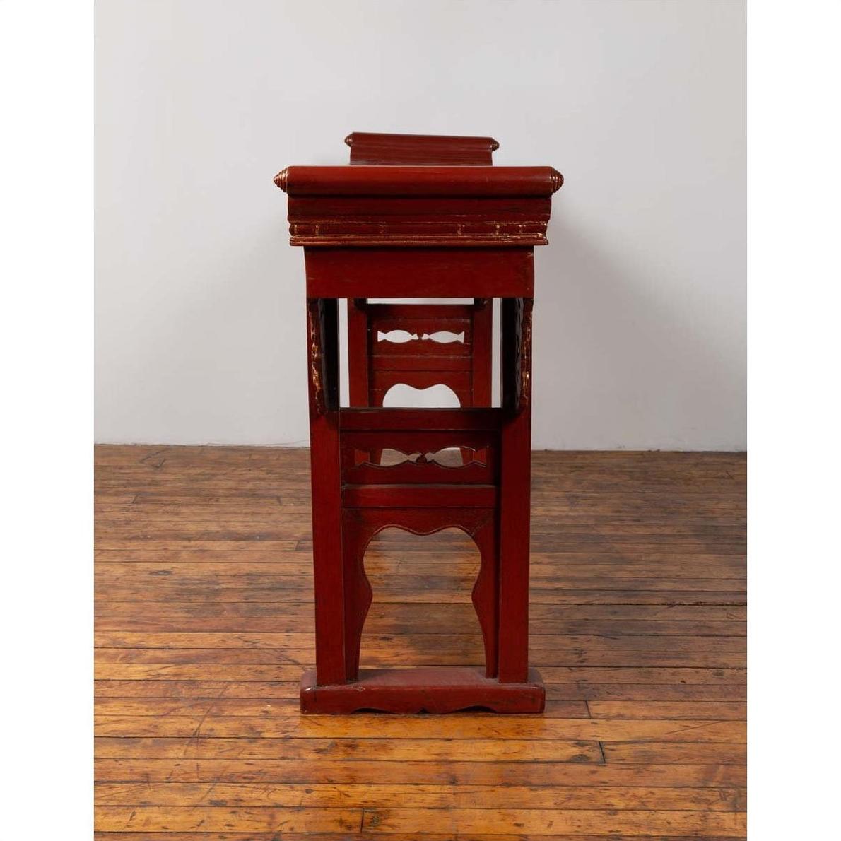 Red lacquered wood console table with carved apron on wood floor against white wall indoors.