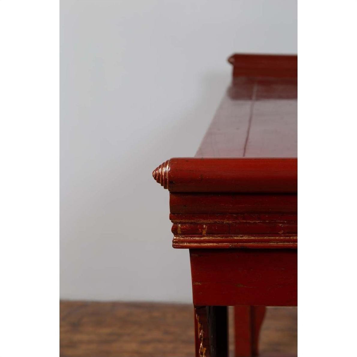 Close-up of red lacquered wood console table with carved apron detailing, plain background.
