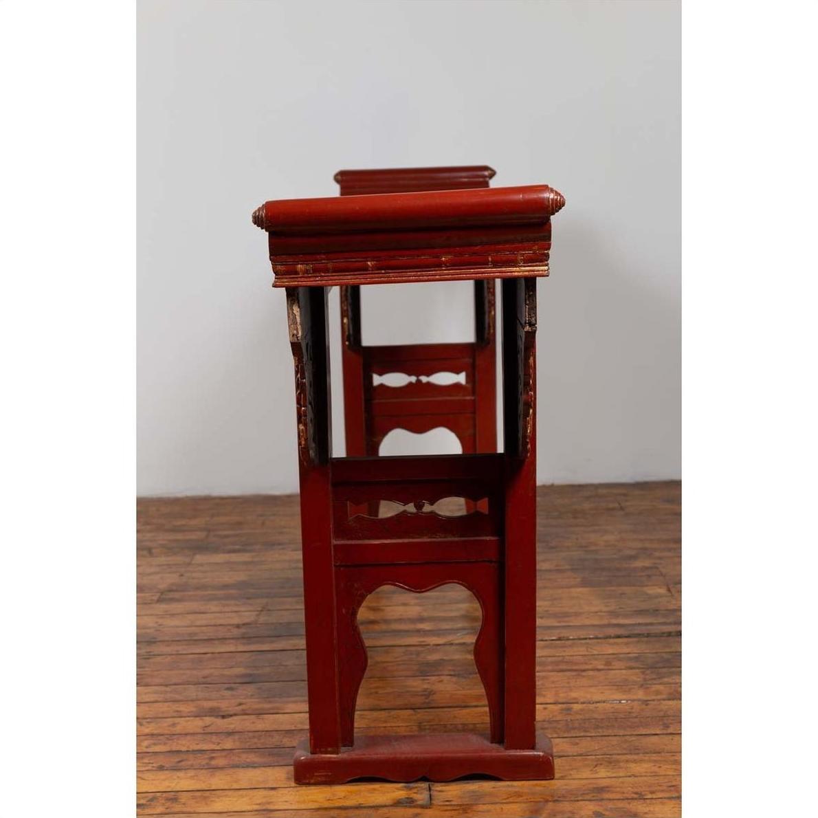 Red lacquered wood console tables with carved aprons, set indoors against a white wall on a wooden floor.