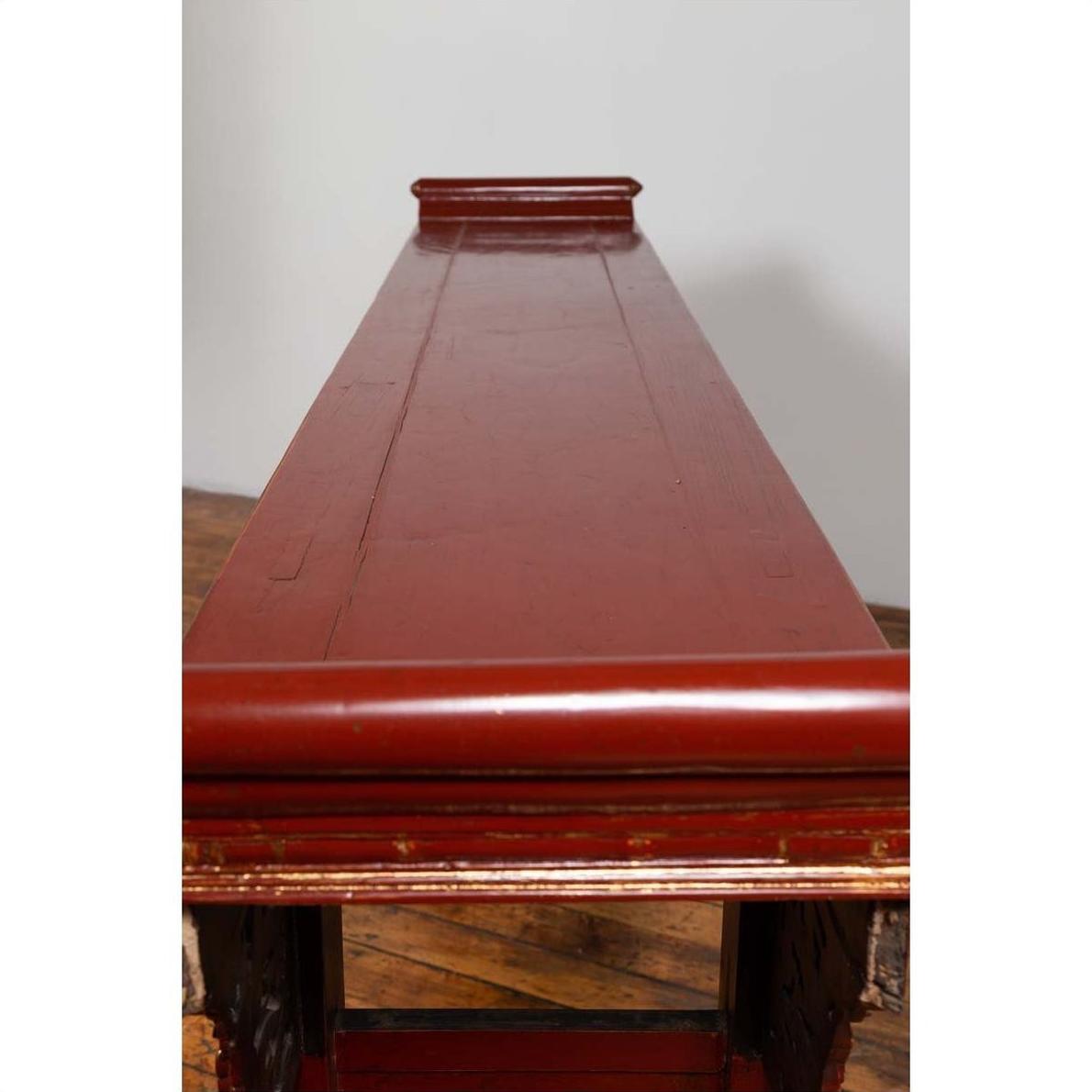 Red lacquered wood console table with carved apron, Ming style, front view on wooden flooring indoors.