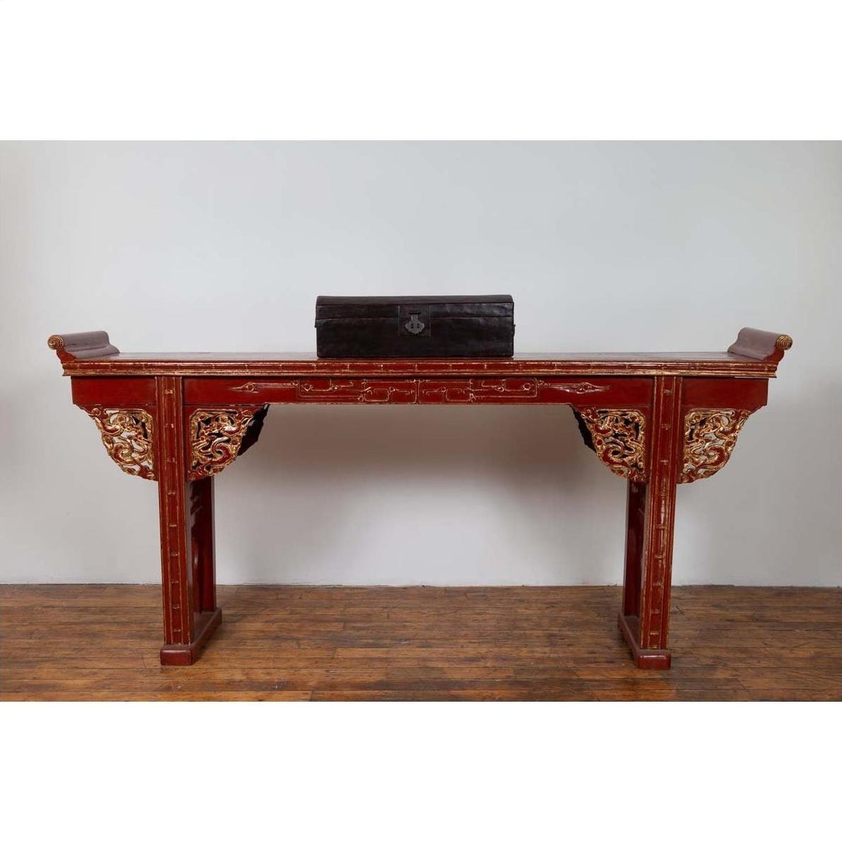 Red lacquered wood console table with carved apron and gold details, displayed indoors against a white wall.