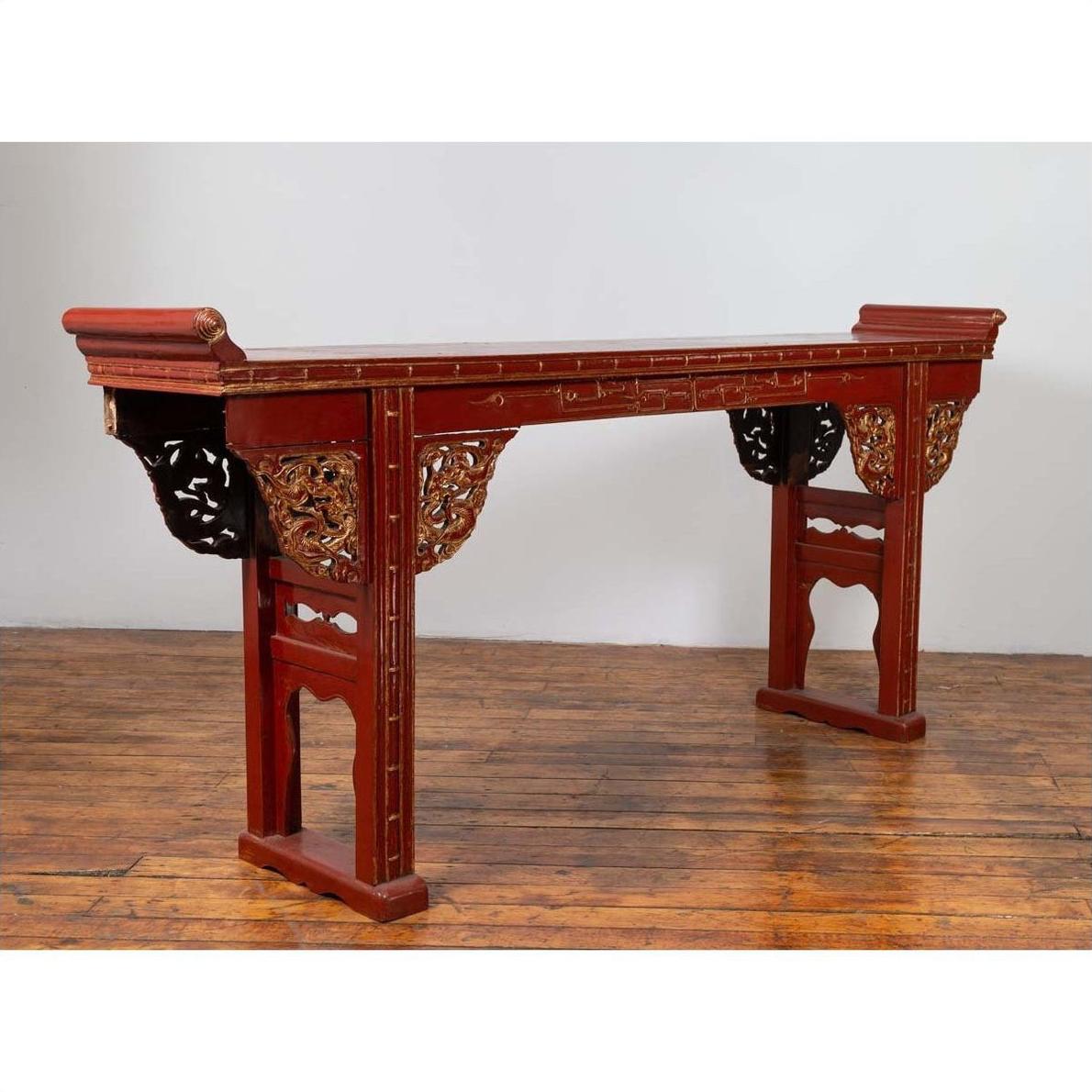 Red lacquered wood console table with carved apron, on wooden floor against a white wall.