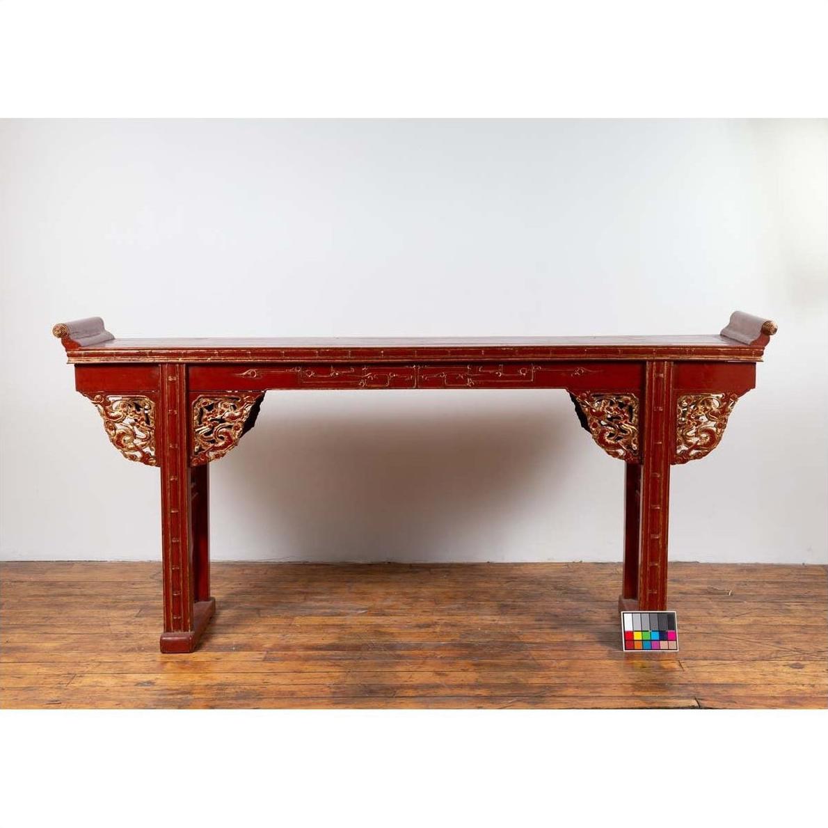 Red lacquered wood console table with carved apron, on wooden floor against a white wall indoors.