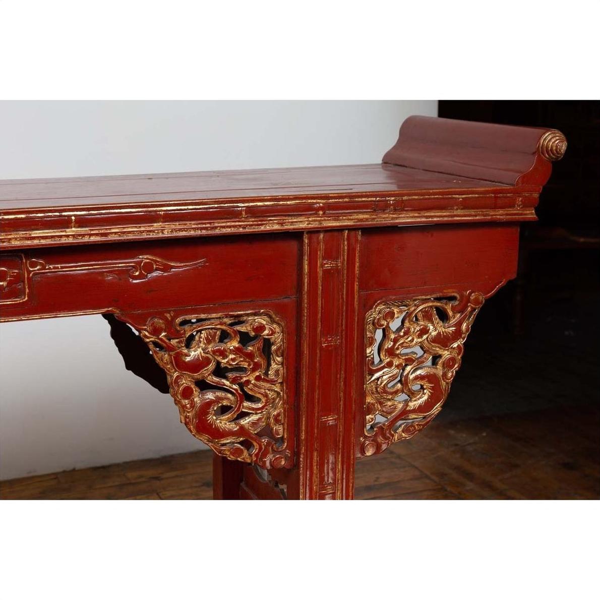 Red lacquered wood console table with carved apron, gold dragon designs, and raised scroll back, indoor setting.