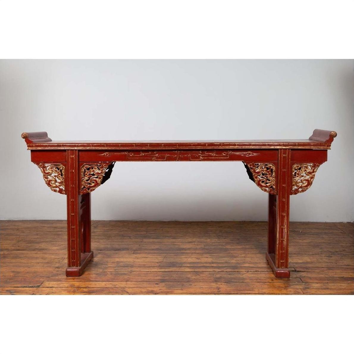 Red lacquered wood console table with carved apron, on wooden floor against plain white wall indoors.