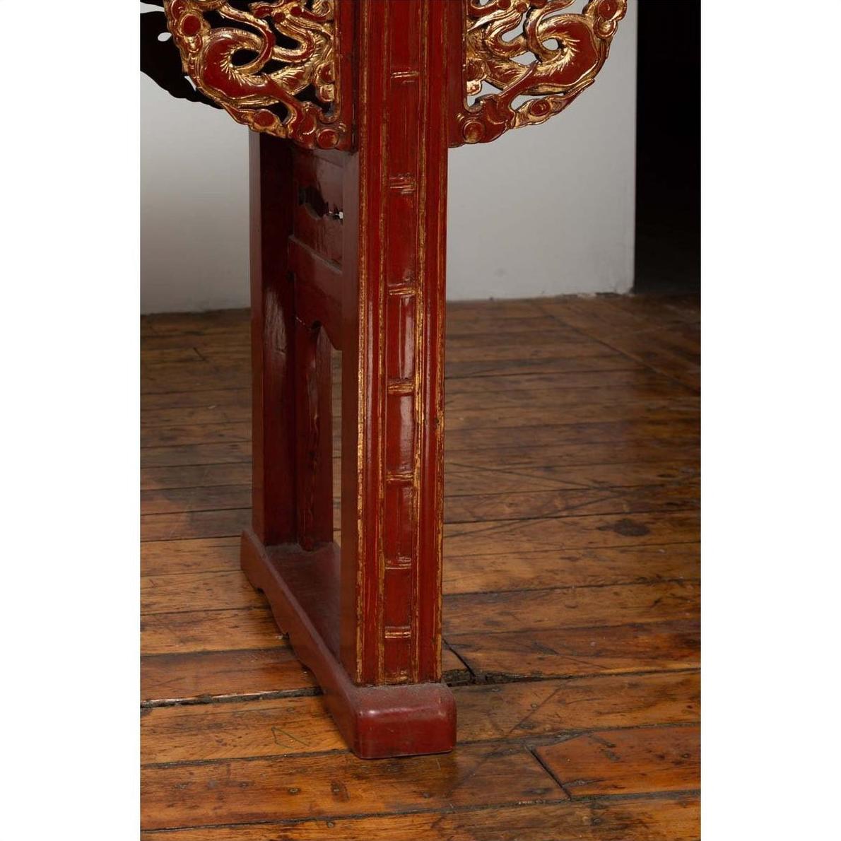 Red lacquered wood console table with carved apron, placed on a worn floor by a white wall in dim lighting indoors.