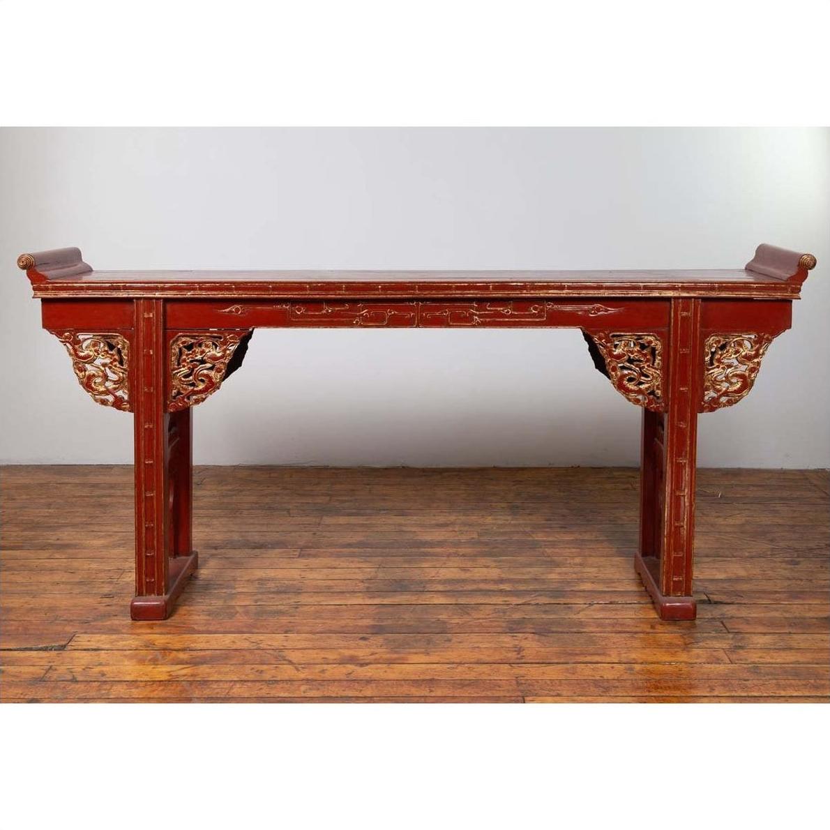 Red lacquered wood console table with carved apron, set on a wooden floor against a plain white wall indoors.