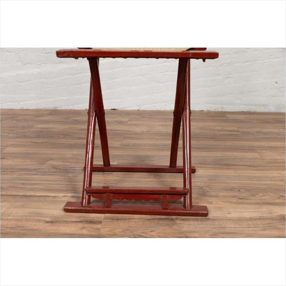 Red lacquered wood folding chair with footrest on wooden floor next to white brick wall.