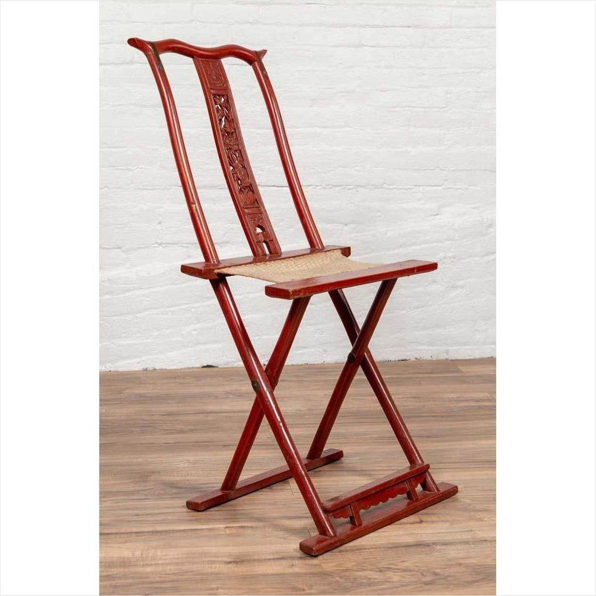 Red lacquered wood folding chair with footrest and fabric, on wooden floor against a white brick wall indoors.