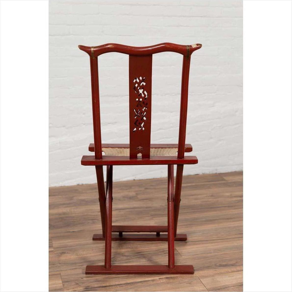 Red lacquered wood folding chair with footrest and fabric, on wooden floor next to white brick wall indoors.