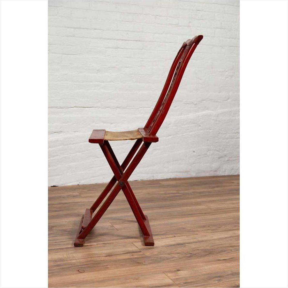 Red lacquered wood folding chair with footrest and fabric, placed indoors against a white brick wall.