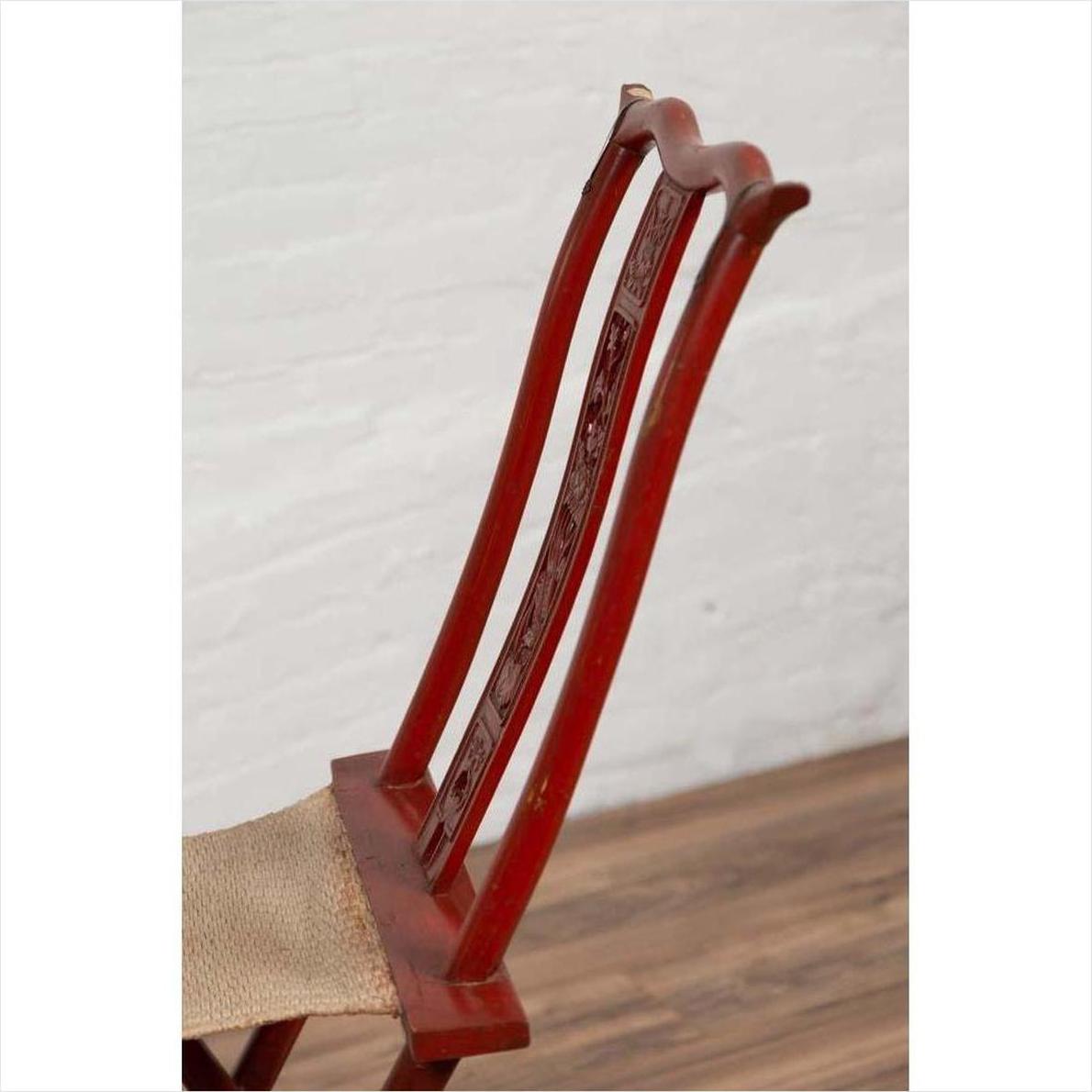 Close-up of a red lacquered wooden folding travel chair with footrest and fabric, indoors against a white wall.