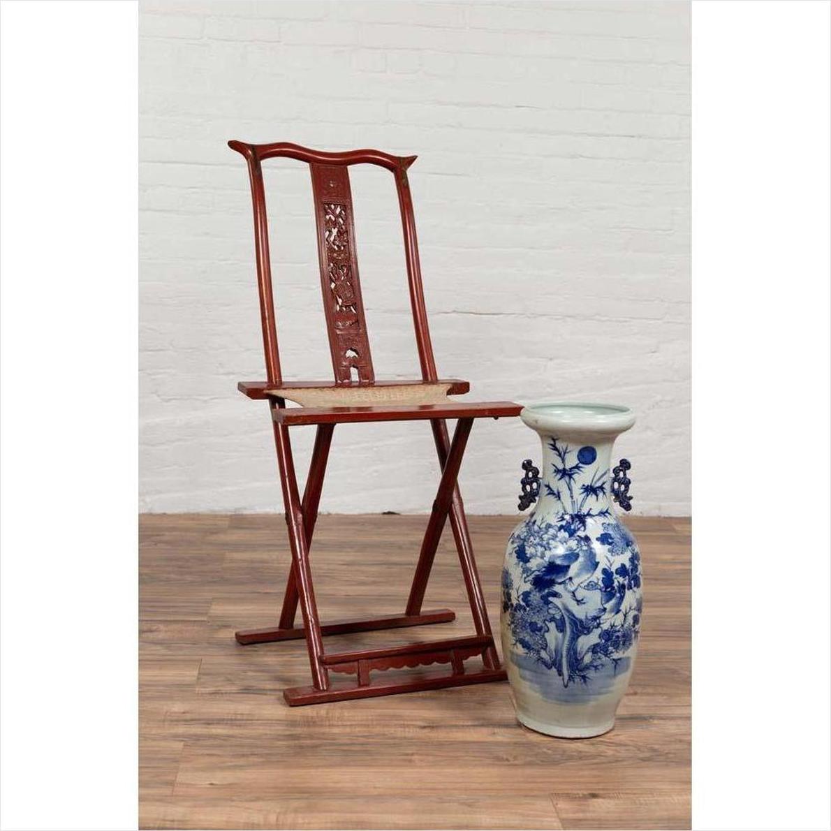 Red lacquered wood folding traveler's chair with footrest next to blue and white vase, indoor setting.