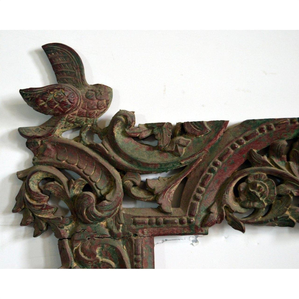Close-up of an antique hand-carved wooden mirror frame with ornate corners, perched bird, floral motifs, green and red hues.
