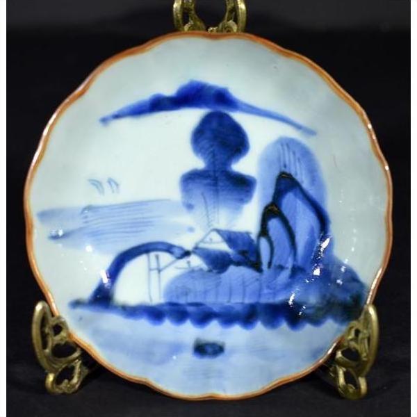 Blue and white hand painted Chinese porcelain bowl with landscape and figures, displayed on gold stand in moody light.