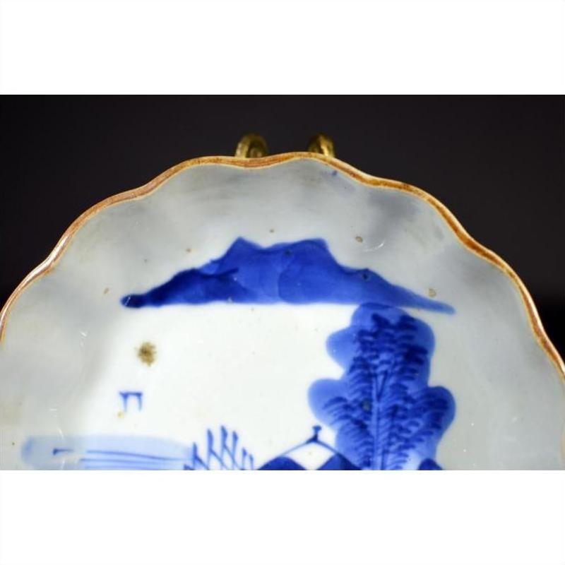 Close-up of blue and white porcelain plate with scalloped edge, hand-painted landscape design, and gold trim.