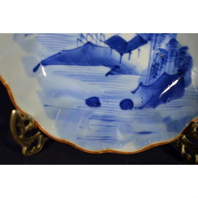Blue and white porcelain plate with hand-painted landscape, scalloped gold-edged rim, displayed on metal stand.