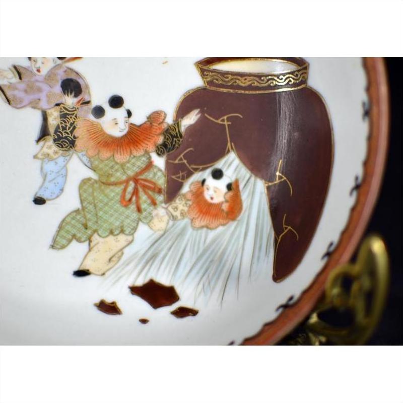 Close-up of antique hand painted Japanese Kutani porcelain plate with green-clad figure aiding another from gold-trimmed jar.