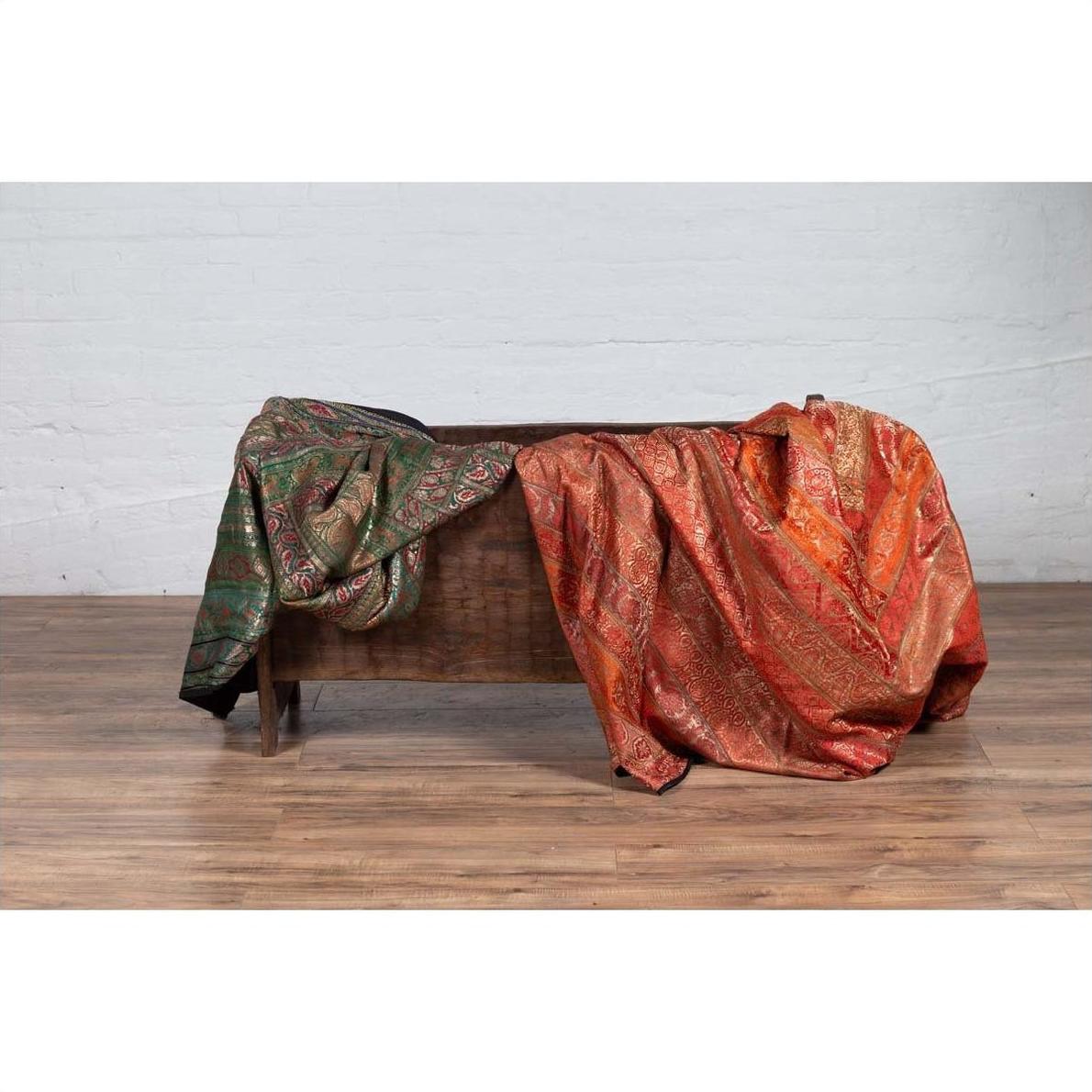 Green and red ornate fabric sarees draped over antique wooden planter on wooden floor, white wall background.